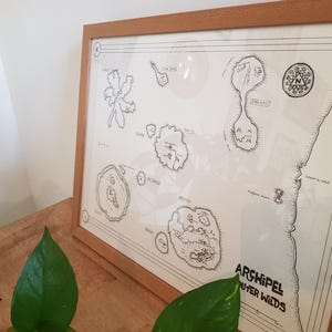 May include: A framed black and white map of a fictional archipelago called 'Archipel Outer Wilds'. The map shows several islands with names like 'Island of the Lost' and 'The Big One'. The map is surrounded by a wooden frame.