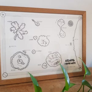 May include: A framed black and white map titled 'Archipel D'Outer Wilds' with a compass rose in the top left corner and a variety of islands and land masses. The map is surrounded by a light brown wood frame.