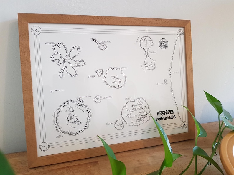 May include: A framed black and white map of the 'Archipel Youtre Wids' with a brown wooden frame. The map shows several islands with names and details. The map is on a light brown surface with a green plant in the foreground.
