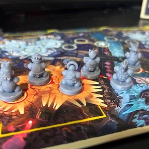 May include: Six gray plastic game pieces with different designs, including a monster, a creature with a crown, and a creature with a hat. The pieces are on a colorful game board with a blue and white design.