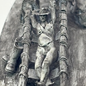 May include: A detailed, gray, 3D printed sculpture of a person hanging from a wooden ladder. The person is wearing a hat and has their arms raised above their head.