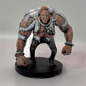 May include: A hand-painted miniature figure of a muscular humanoid with a stern expression. The figure has a tan torso, black pants, and silver accents on the shoulders, wrists, and neck. It stands on a black circular base.