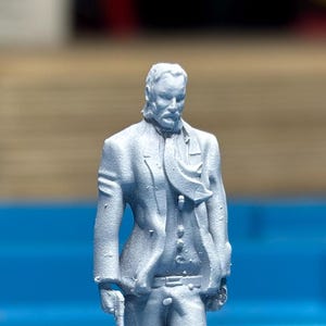 May include: A light gray figurine of a man in a suit, with a long coat, a scarf, and a mustache. The figure is holding a cane. The statue is detailed and appears to be made of a hard material.
