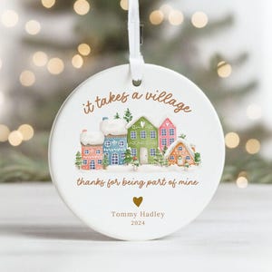 May include: White ceramic Christmas ornament with a village scene and the text "It takes a village thanks for being part of mine."  The ornament also includes the name "Tommy Hadley" and the year "2024".