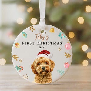 May include: A round, white Christmas ornament with a festive design. It features a portrait of a dog wearing a Santa hat, with the text "Toby's FIRST CHRISTMAS 2024" in elegant script. The ornament is adorned with holiday-themed illustrations.