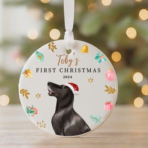 May include: White ceramic Christmas ornament with a black dog wearing a Santa hat. The ornament has the text "Toby's First Christmas 2024" and festive illustrations.