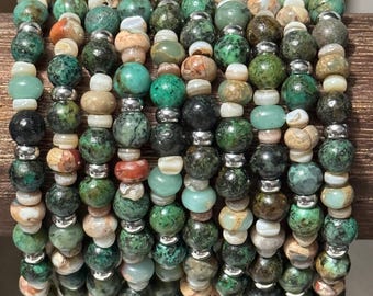 Può includere: Una collezione di braccialetti di perline con una varietà di perline di pietra naturale nei toni del verde, marrone e crema. I distanziatori color argento si alternano alle perline, creando un accessorio strutturato e colorato.