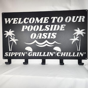 May include: Black metal sign with white text that reads "Welcome to our Poolside Oasis Sippin' Grillin' Chillin'" with palm trees and waves. The sign has five hooks.