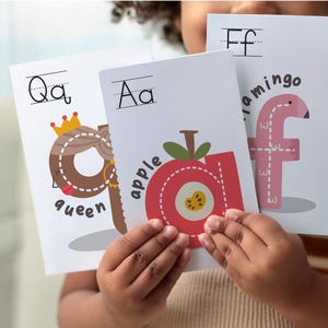 Op de afbeelding: Een kind houdt drie alfabetkaarten vast. De kaarten tonen de letters Q, A en F, met illustraties van een koningin, appel en flamingo. De kaarten zijn wit met kleurrijke illustraties en tekst.