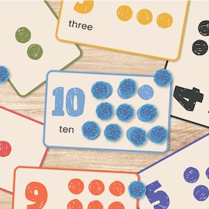 May include: Educational flashcards with numbers and corresponding quantities. The cards feature numbers from one to ten, written in a playful font, and are accompanied by colorful dots or pom-poms. The background is a light wood grain.