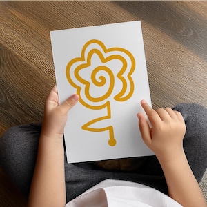 Könnte beinhalten: Eine Person hält eine weiße Karte mit einer gelben Blumenillustration. Das Blumendesign ist abstrakt, mit einem spiralförmigen Zentrum und einem Stiel. Die Person trägt eine graue Hose und sitzt auf einem Holzboden.