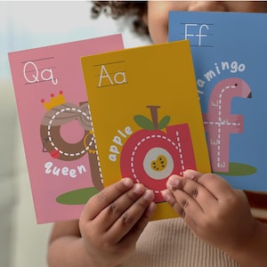 May include: Three colorful alphabet flashcards are held in front of a person. The cards feature the letters Q, A, and F, with corresponding illustrations of a queen, an apple, and a flamingo. The cards are pink, yellow, and blue.