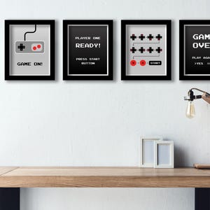 Könnte beinhalten: Drei schwarze gerahmte Drucke mit weißen Hintergründen und schwarzem Text. Der erste Druck zeigt einen grauen Videospielcontroller mit dem Text "GAME ON!" Der zweite Druck zeigt den Text "PLAYER ONE READY!" mit dem Text "PRESS START BUTTON" darunter. Der dritte Druck zeigt ein Diagramm eines Videospielcontrollers mit dem Text "GAME OVER" und "PLAY AGAIN? YES NO" darunter.