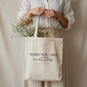 May include: A beige canvas tote bag with the words "WHO YOU ARE matters" printed in black. The bag is filled with white flowers and held by a person. The Small Elephant | EST 2023 is printed at the bottom.