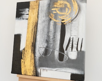 Könnte beinhalten: Abstrakte Leinwandkunst mit einer Farbpalette aus Gold, Schwarz, Weiß und Grau. Das Kunstwerk zeigt einen vertikalen Goldstreifen, schwarze Formen und ein kreisförmiges Golddesign. Die Leinwand wird auf einer Holzstaffelei ausgestellt.