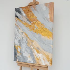May include: Abstract painting on canvas with a palette of gold, gray, and beige. The artwork features textured brushstrokes and is displayed on a wooden easel. The composition has a diagonal flow, creating a dynamic visual effect.