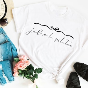 May include: A white short-sleeved t-shirt featuring the black cursive text j'adore le pilates centered on the chest, accompanied by a black line drawing of a decorative bow above the lettering. The shirt is made of a plain white fabric.