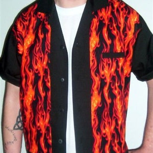 Puede incluir: Camisa negra con botones y mangas cortas con diseño de llamas. La camisa presenta cuello, bolsillo y mangas negros, con un vibrante patrón de llamas naranja y rojo en los paneles delanteros.