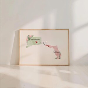 May include: A framed wall art print featuring a light green map of Connecticut and a light pink map of Florida. The states are connected by a dashed black line with two red hearts and a heart-shaped loop. Floral accents decorate the edges.