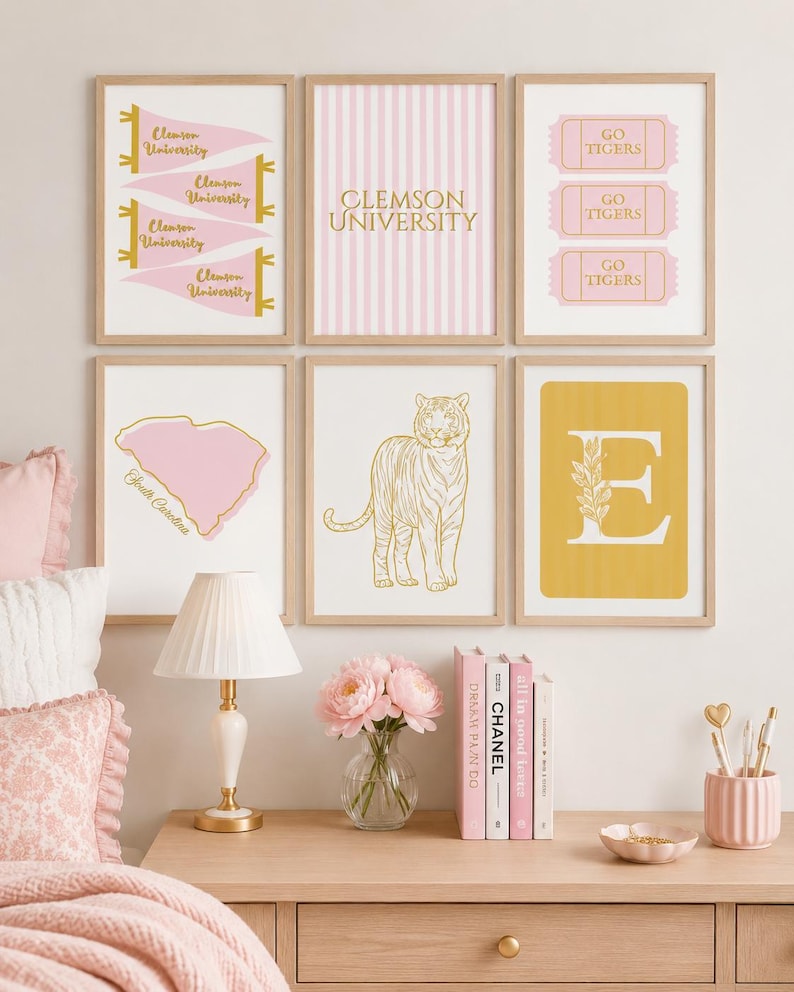 May include: A gallery wall set of six framed prints featuring Clemson University themes in pink and gold. Designs include pennants, striped text, ticket stubs, a map of South Carolina, a tiger illustration, and a large letter E with floral detail.