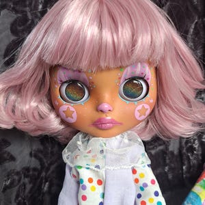 Può includere: Una bambola con i capelli rosa e una camicia bianca con pois colorati. La bambola ha un trucco per gli occhi arcobaleno e un naso rosa. La bambola indossa un colletto di pizzo bianco.