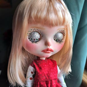 May include: A Blythe doll with light blonde hair and glittery silver eye makeup with red and green accents. The doll has rosy cheeks and red lips, wearing a red dress with a white and green floral patterned top.