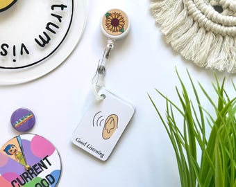 May include: A white retractable badge reel with a sunflower design and a white ID card that says "Good Listening" with an illustration of an ear. Other items include a pin and a mood wheel.