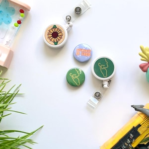 Puede incluir: Cuatro botones diferentes con fondo blanco y diseños coloridos. Los botones muestran un girasol, un contorno verde de una persona, un botón azul con el texto "La amabilidad es gratis", y un botón verde con un contorno blanco de una persona. Los botones están unidos a carretes retráctiles de identificación blancos.