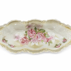 May include: An oval-shaped ceramic dish with a scalloped, gold-trimmed edge. The center features a painted cluster of pink roses with green leaves, while smaller rose motifs are positioned on the left and right sides against a cream background.