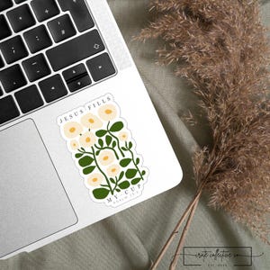 May include: A white sticker with the text "JESUS FILLS MY CUP" and "PSALM 23:5" features a floral design with cream-colored flowers and green leaves. The sticker is on a laptop, with a keyboard visible. Dried plants are in the background.
