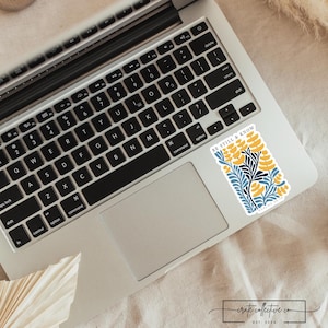 Puede incluir: Una pegatina con el texto "BE STILL & KNOW" y un diseño floral en amarillo y azul está colocada en un ordenador portátil. El portátil está abierto, mostrando su teclado y su panel táctil. La pegatina tiene un fondo blanco.