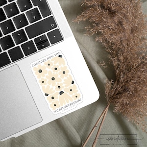 Puede incluir: Una pegatina rectangular con un diseño floral con flores de color crema con centros negros. La pegatina tiene el texto "CONSIDER HOW THE WILDFLOWERS GROW" y "LUKE 12:27". La pegatina está en un portátil.