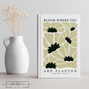 May include: A white book with the text "BLOOM WHERE YOU ARE PLANTED" and a floral design in black and olive green. A white vase with dried flowers is to the left. The book's design is a modern, minimalist style.