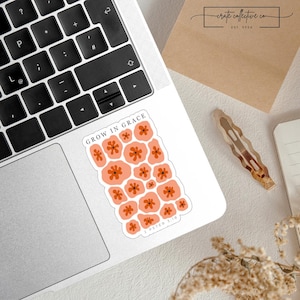 Puede incluir: Una pegatina con el texto "GROW IN GRACE" y un diseño floral en tonos melocotón y naranja. La pegatina está sobre un portátil, con el teclado a la vista. La pegatina también tiene el texto "2 PETER 3:18".