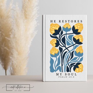 Puede incluir: Un libro blanco con el texto "HE RESTORES MY SOUL" y el Salmo 23:3. La portada presenta un diseño floral con flores amarillas y azules. El libro está sobre una superficie blanca con plantas secas en el fondo.