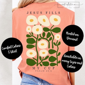 Puede incluir: Camiseta Comfort Colors color coral con diseño floral y el texto "JESUS FILLS MY CUP PSALM 23:5". El gráfico presenta flores blancas con centros amarillos y hojas verdes. Disponible en muchas tallas y colores.
