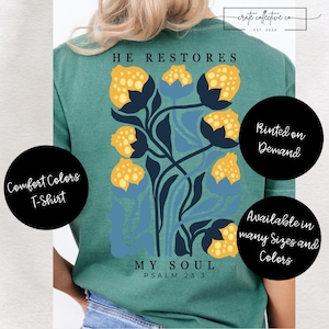 Puede incluir: Camiseta Comfort Colors de color verde azulado con un diseño floral en amarillo y azul. El texto "HE RESTORES MY SOUL" está impreso en la camiseta, junto con "PSALM 23:3". Disponible en muchas tallas y colores.