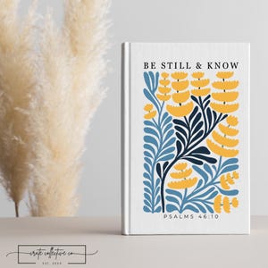 Puede incluir: Un diario blanco con el texto "BE STILL & KNOW" y "PSALMS 46:10". La portada presenta un diseño floral en amarillo, azul y marino. El diario está de pie.