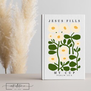 Puede incluir: Un libro blanco con el texto "JESUS FILLS MY CUP PSALM 23:5" y un diseño floral en amarillo y verde. El libro está de pie. El fondo incluye hierba ornamental seca.