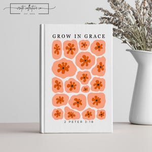 以下が含まれることがあります： 「GROW IN GRACE」と「2 PETER 3:18」の文字が入った白い本の表紙。オレンジ色の中心を持つ、桃色の花のパターンが特徴です。本は、ドライフラワーの花瓶と一緒にテーブルの上に展示されています。