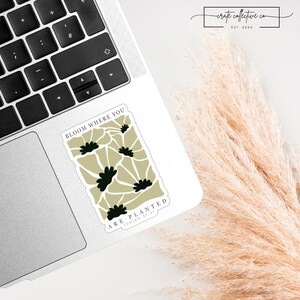 Puede incluir: Una pegatina rectangular con el texto "BLOOM WHERE YOU ARE PLANTED" y un diseño floral en negro y verde oliva. La pegatina está sobre un portátil, con un teclado visible. La esquina superior derecha presenta el texto "-crate collective co. EST. 2024".