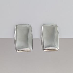 May include: A pair of silver rectangular stud earrings. Each earring has a slightly rounded shape with a smooth, reflective surface. The earrings are displayed against a white and grey background.