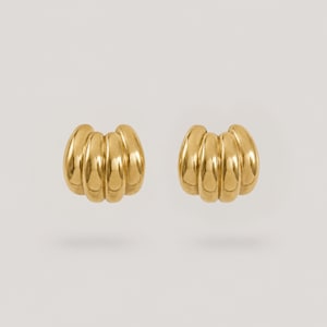 Könnte beinhalten: Ein Paar goldfarbene Ohrstecker mit einem abgerundeten, gerippten, croissantartigen Design und einer polierten, reflektierenden Oberfläche. Die Ohrringe haben eine geschwungene, mehrteilige Form, die ein dimensionales Aussehen erzeugt.