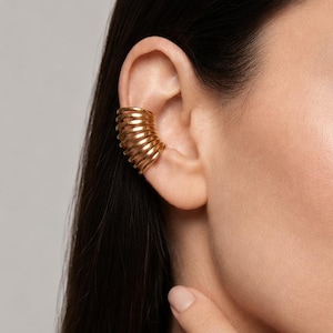 May include: A gold-toned ear cuff featuring a series of stacked, rounded wire bands that wrap around the outer edge of the ear cartilage. The jewellery piece has a polished, metallic finish and a continuous, ribbed, coil-like structure.