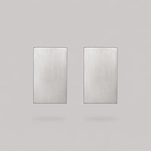 Puede incluir: Un par de pendientes rectangulares con un acabado de metal plateado cepillado. Cada pendiente tiene una orientación vertical plana con bordes rectos y limpios, y una textura de superficie uniforme y lisa que refleja la luz de manera pareja.