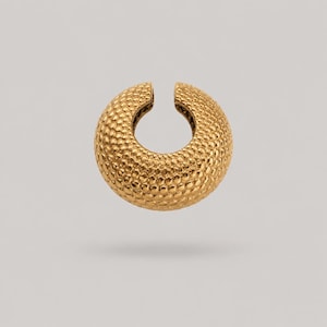 May include: A gold-toned ear cuff featuring a circular, open-ended design with a textured, dimpled surface. The piece has a rounded, thick profile and a polished finish, creating a consistent pattern of small, raised bumps across the entire exterior.