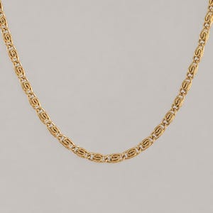 Puede incluir: Un collar de cadena de metal en tono dorado que presenta un patrón repetitivo de eslabones entrelazados con forma de S estilizada. La cadena tiene un acabado pulido y un diseño uniforme y consistente en toda su longitud, formando una curva.