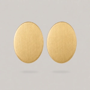May include: A pair of gold-toned earrings featuring a simple oval shape with a brushed, matte finish. The surface of each earring displays a subtle vertical texture, creating a uniform appearance across both pieces in this set of jewellery.