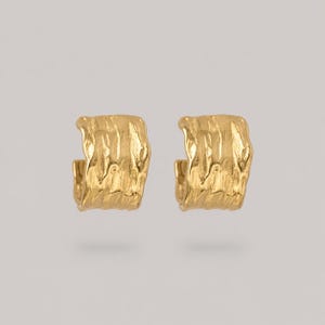 May include: A pair of gold hoop earrings featuring a textured, irregular surface with vertical ridges. The earrings have a wide, open-ended design with a polished finish that reflects light across the uneven, organic-looking metal surface.