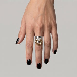 Op de afbeelding: Een brede zilveren ring met een centraal goudkleurig cirkelvormig element. De gouden cirkel is geaccentueerd met een enkele, heldere, vierkant geslepen edelsteen aan de bovenzijde. De ring heeft een gepolijste afwerking en een dik profiel.
